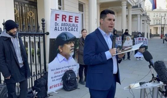 Burgon speaks to the press , with Ali Mushaima's picture in the back