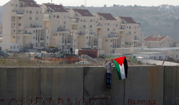 A man places the Palestinian flag on the controversial Israeli separation wall, with Israeli settlements showing in the background. (Photo: Reuters/Ammar Awad]