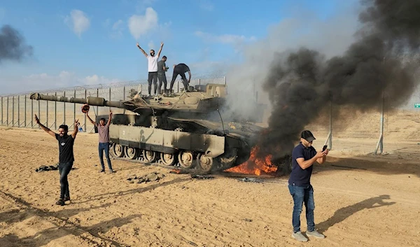 Palestinians celebrate by a destroyed Israeli tank at the Gaza Strip fence east of Khan Yunis, occupied Palestine, October 7, 2023 (AP)