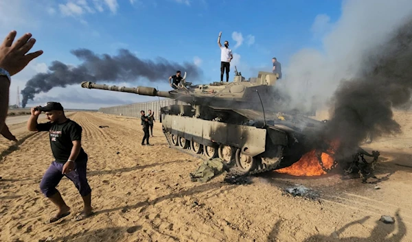 Palestinians celebrate by a destroyed Israeli tank at the Gaza Strip separation wall east of Khan Yunis, occupied Palestine, October 7, 2023 (AP)