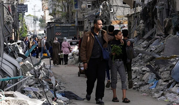 Palestinians walk by buildings destroyed in the Israeli bombardment of the Gaza Strip in Nusseirat refugee camp, central Gaza Strip, Saturday, Nov. 25, 2023 (AP)