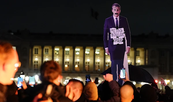 Demonstrators hold a placard depicting French President Emmanuel Macron that reads, "metro, work, grave" during a protest in Paris, March 17, 2023 (AP)