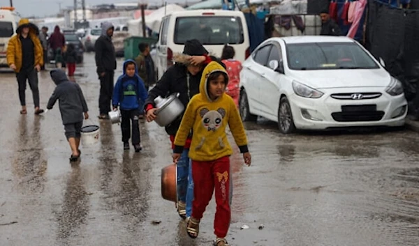 Forcibly displaced Palestinian children walk in a puddle in rainy weather to get food rations at a makeshift tent camp in Rafah in the southern Gaza Strip on February 2, 2024, amid ongoing Israeli genocide. (AFP)