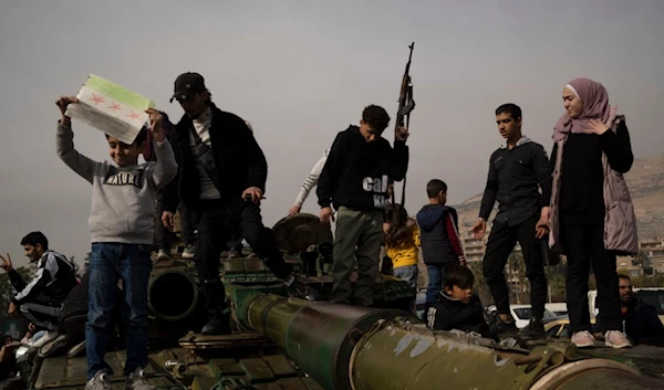 A youth holding a rifle borrowed from a Syrian opposition fighter poses for a picture on the top of a government forces tank that was left on a street, at the Umayyad Square in Damascus, Syria, Wednesday, December 11, 2024 (AP)