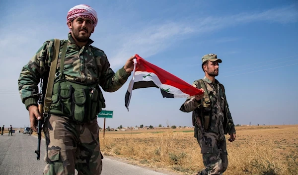 Syrian government forces carry a national flag as they walk at a checkpoint near the town of Tal Tamr, al-Hasakah, north Syria, on Oct. 22, 2019.  (AP)