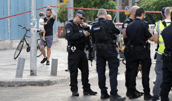 An Israeli settlers walks past Israeli occupation forces investigating the scene of a deadly explosion in Tel Aviv, occupied Palestine, July 19, 2024 (AP)