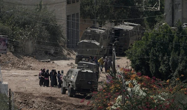 Palestinians stand in line next to Israeli armoured vehicles during a military operation in the West Bank Jenin refugee camp, Saturday, Aug. 31, 2024. (AP Photo/Majdi Mohammed)